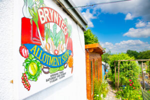 picture of a board for Brynteg Allotment shop with a shed and plants in the background