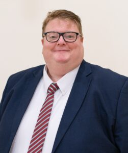 Head shot of a gentleman with ginger hair and glasses. Wearing a suit.